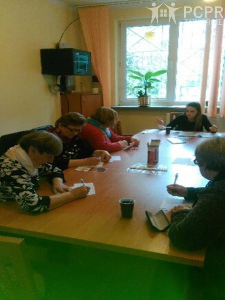 Zdjęcie: Uczestnicy i uczestniczki siedząc przy stole słuchają wypowiedzi osoby prowadzącej zajęcia